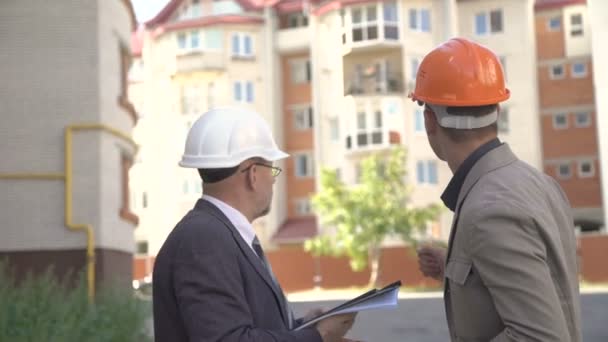 Dos hombres de negocios en casco discutiendo esquema de construcción cerca de construye. 4K — Vídeos de Stock