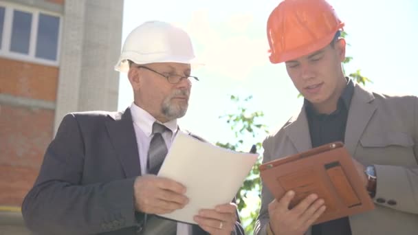 Dois trabalhadores discutindo esquema de construção usando tablet perto constrói. 4K — Vídeo de Stock