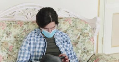 Adam kanepede dinlenirken telefon kullanıyor, maskesini indiriyor ve yalnızken yorgun düşüyor.