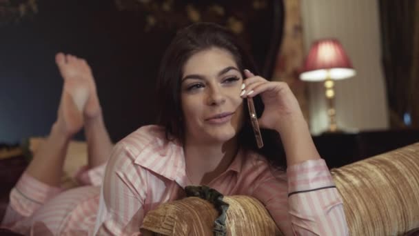 Enigmatisk brunett som ligger på sängen och pratar i telefon — Stockvideo