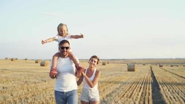 Caminhada de família alegre no campo, menina em ombros de pais mostra polegar — Vídeo de Stock