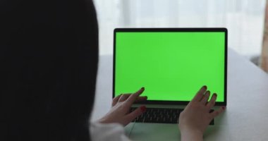 Dizüstü bilgisayarda yazan ve penceredeki masada yeşil ekran olan bir kadın.