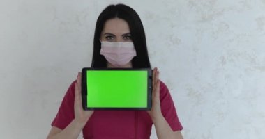 Nurse in medical mask holds tablet and shows screen at camera on background