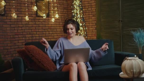 Chica con estilo utiliza un ordenador portátil, escucha música en los auriculares, canta y baila — Vídeo de stock