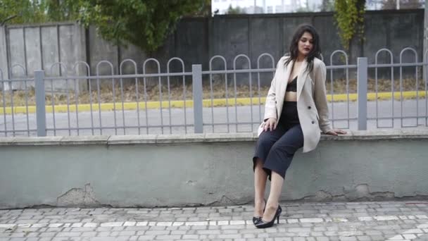 Elegancka brunetka w stylowych ubraniach siedząca przy ogrodzeniu w pozach z radością na twarzy — Wideo stockowe