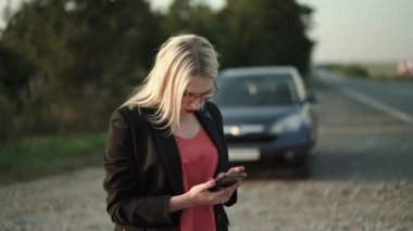 Kafası karışmış bir kadın telefon ekranında takla atar ve etrafa bozuk arabaya bakar.