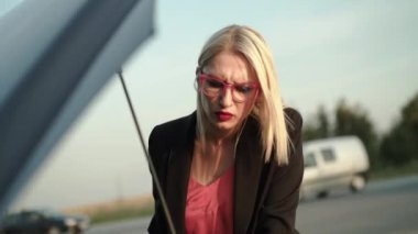 Gözlüklü kafası karışmış bir sarışın bozuk bir arabanın kaputunun altına bakar ve yana bakar.