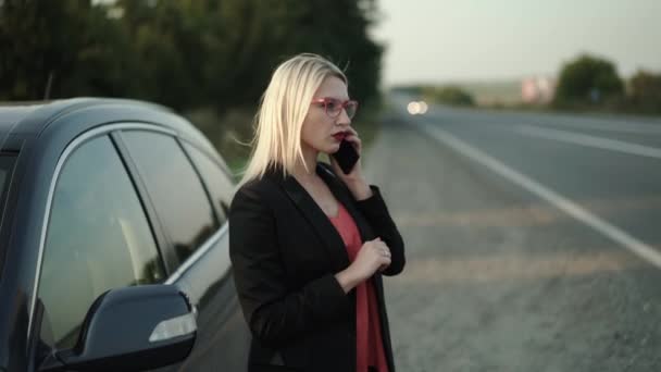 Μπερδεμένη ξανθιά με γυαλιά μιλάει σε ένα smartphone συναισθηματικά σε ένα σπασμένο αυτοκίνητο — Αρχείο Βίντεο