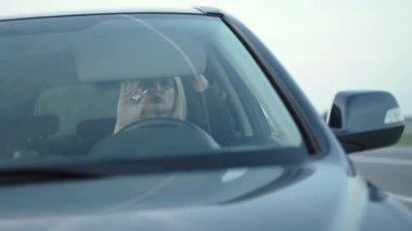 Sarışının arabadaki aynaya kırmızı arpacığı uyguladığı ön camdan bak