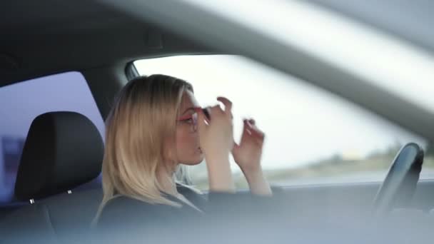 Blonda în ochelari corectează părul și aplică pomada roșie în oglindă în mașină — Videoclip de stoc