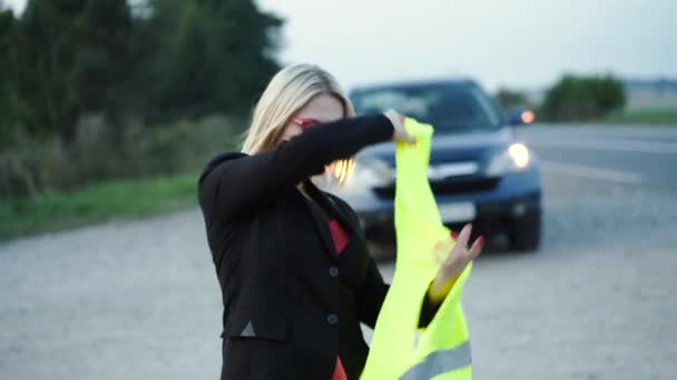 Mladá dáma nosí reflexní bezpečnostní vestu a dívá se do kamery na rozbité auto — Stock video