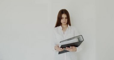 Genç kız elinde siyah klasörle sarılıp beyaz arka plandaki kameraya bakıyor.