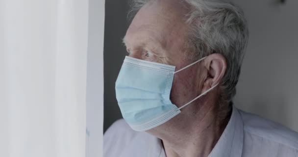 Lonely grandfather in medical mask looking through window, leaning to wall — Stock Video