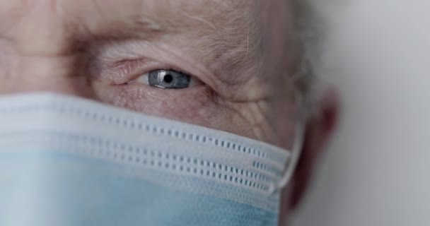 Μακρο-πρόσωπο μέρος του γαλανομάτη ηλικιωμένος με μάσκα κοιτάζοντας κάμερα στο παρασκήνιο — Αρχείο Βίντεο