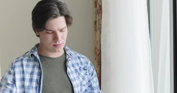 Mladý muž na izolaci doma nosí masku a dívá se z okna — Stock video