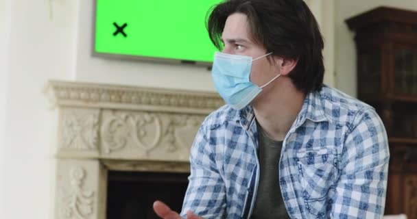 Zenuwachtige man met masker geïsoleerd thuis, denkt en katheert hoofd op tv plasma — Stockvideo