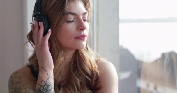 Kobieta w słuchawkach, dotykając nauszników i uśmiechając się do okna — Wideo stockowe