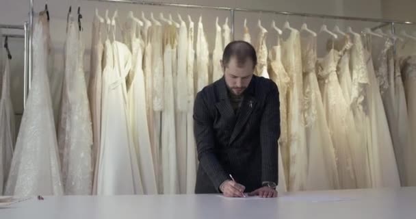 Man ontwerper tekent jurk schets op een bureau in bruiloft atelier — Stockvideo