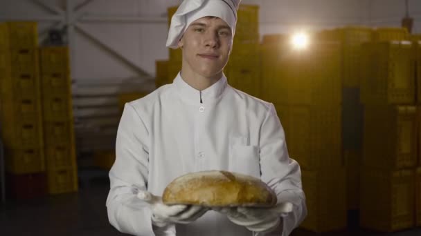 Gelukkig bakker in uniform presenteert sluiten op camera een vers gebakken brood — Stockvideo