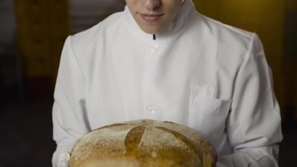 Baker em uniforme cheira um pão, tem um prazer e sorriso na câmera — Vídeo de Stock