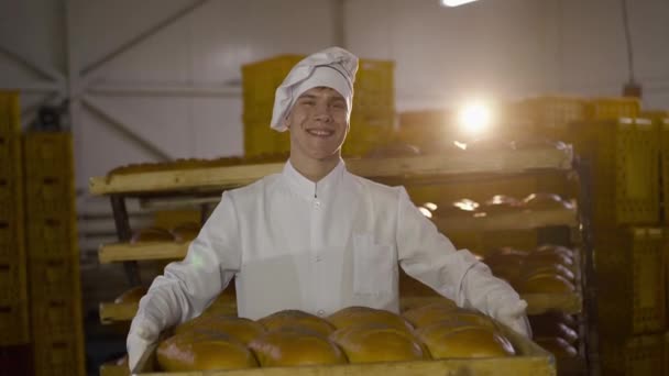 Jeune boulanger en uniforme présente un plateau de pain de blé à la caméra dans la boulangerie — Video