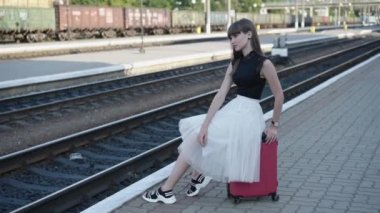 Siyah beyaz elbiseli güzel bir kız demiryolunda sabit bir poz içinde bavulun üzerinde oturuyor.