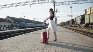 Elbise giymiş çekici bir model, tren istasyonunda bavulla poz veriyor.
