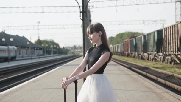 Bedårande modell i svart-vit klänning poser med röd resväska på järnvägsplattform — Stockvideo