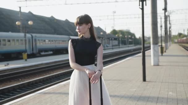 Гламурная модель в черно-белом платье позирует с красным чемоданом на железнодорожной платформе — стоковое видео