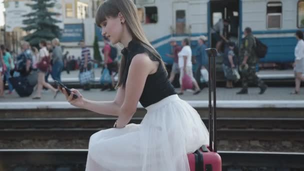 Chica feliz se sienta en la maleta, habla por teléfono con sonrisa y las olas de la mano — Vídeos de Stock