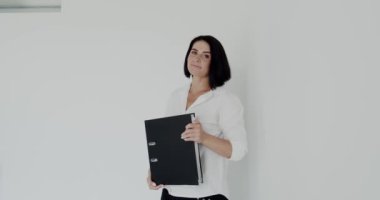 Elinde siyah klasör olan mutlu öğretmen ofiste kameraya gülümsüyor.