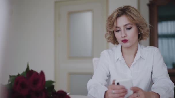 Portret troskliwej pani wykorzystuje telefon przy stole z bukietem róż w domu — Wideo stockowe