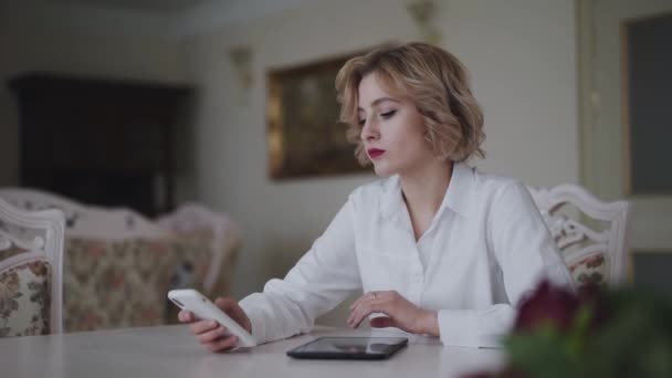 Mujer feliz utiliza teléfono inteligente y tableta en la mesa en la sala de luz — Vídeo de stock
