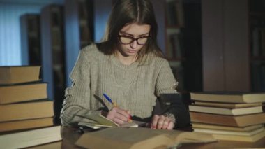 Genç kız kitaplardan bilgi alıyor, kenara dönüyor ve kütüphanede gülümsüyor.