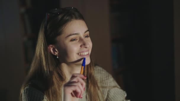 Chica feliz con la pluma en la mano linda sonriendo a la cámara en el escritorio en la biblioteca tenue — Vídeo de stock