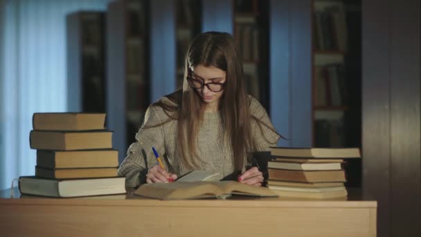 Menina bonita de óculos lê um livro na mesa na biblioteca, cruza as mãos e sorri — Vídeo de Stock