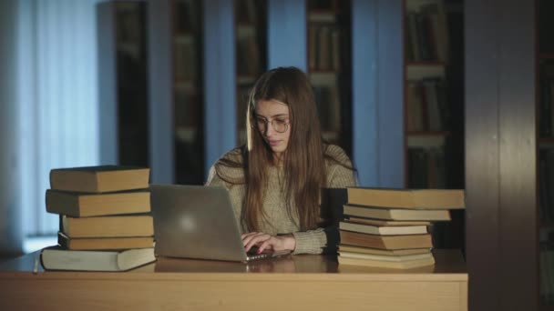 Dziewczyna pracująca z laptopem przy biurku z książkami i uśmiechnięta w bibliotece późno — Wideo stockowe