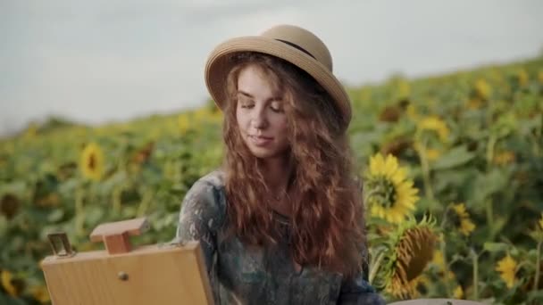 Длинноволосая женщина рисует акварелью среди подсолнухов в ветреный день — стоковое видео