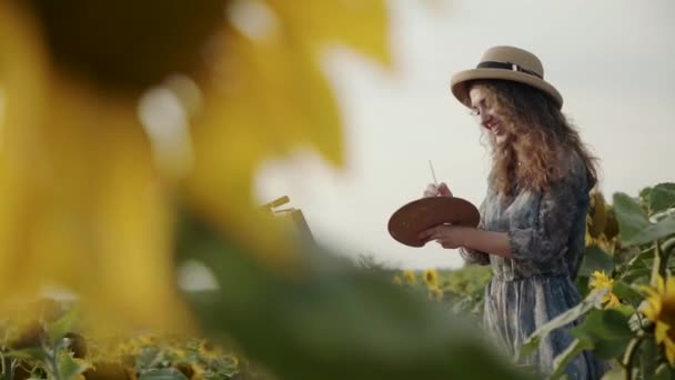 Uitzicht op zonnebloem van vrolijk meisje in hoed met blazen lang haar tijdens het schilderen — Stockvideo