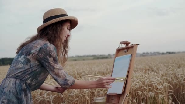 Pretty paintress in hat and dress painting landscape on canvas among wheat field — Stock Video