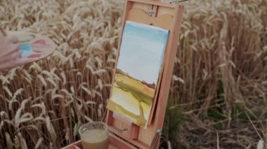 Dişi el, altın buğday arasında fırça ve suluboya ile bir manzara çiziyor.