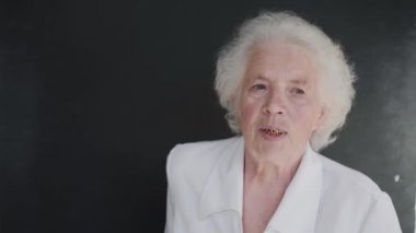 Beyaz ceketli gri saçlı kadının portresi arka planda kameraya bakıyor.