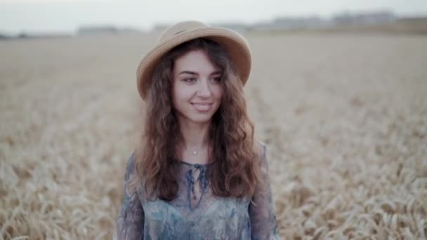 Chica alegre en vestido y sombrero camina en el campo de trigo y sonríe a la cámara — Vídeos de Stock