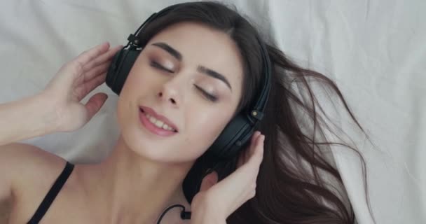 Menina feliz ouvindo música na cama, flerta na câmera, torce o fio dos fones de ouvido — Vídeo de Stock