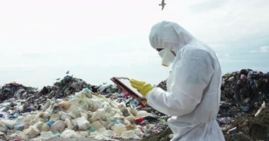 Koruyucu giysili ve gaz maskeli bir adam çöp sahasında tabletine not alıyor.
