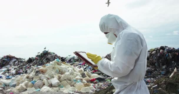 Mężczyzna w kombinezonie ochronnym i masce gazowej, notowany w tablecie na wysypisku śmieci. — Wideo stockowe