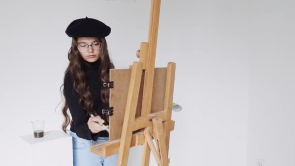 Jonge vrouw kunstenaar in baret schilderij schilderij op doek in kunst atelier — Stockvideo