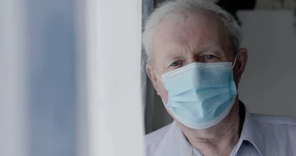 Portrait of sad grandfather in medical mask looks at camera anxiously at home Stock Picture