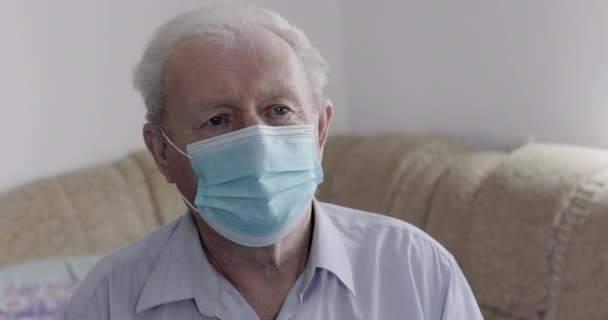 Сумний старший чоловік у медичній масці сидить на дивані і дивиться крізь вікно — стокове відео