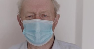 Tıbbi maske takmış yakışıklı bir adam kameraya ciddi ciddi bakıyor.